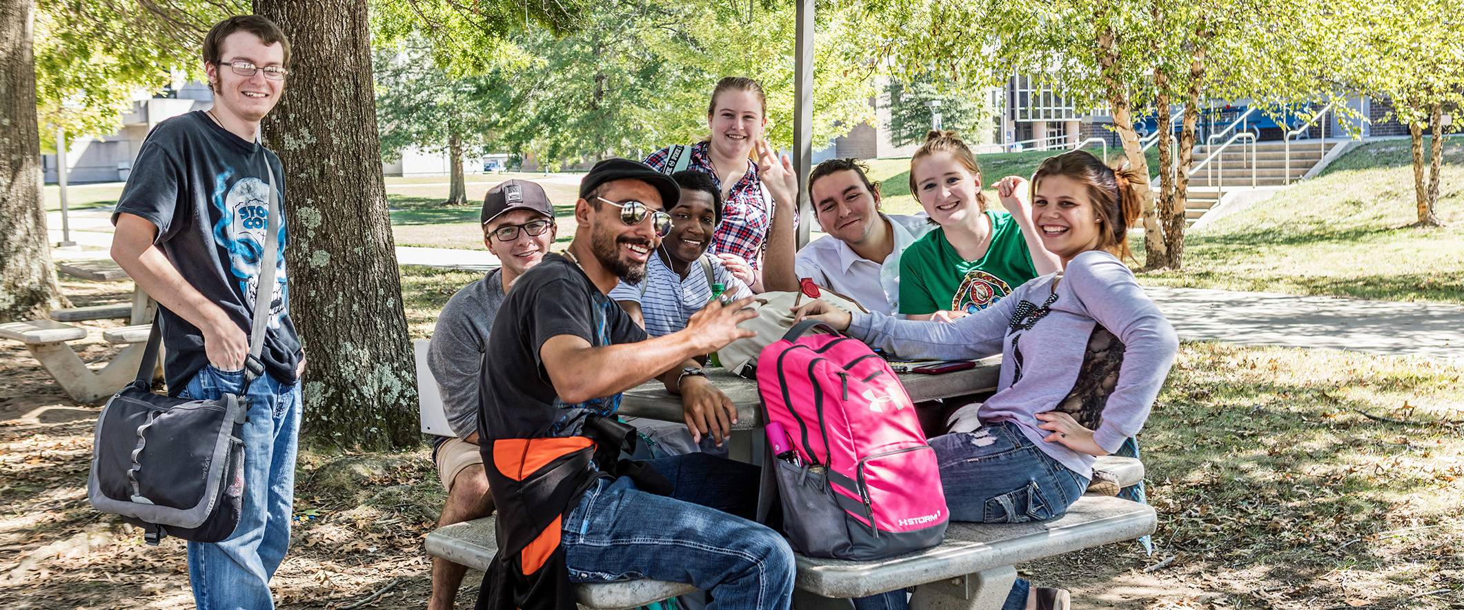 Group of students sitting at a picnic table under the shade of the campus trees
