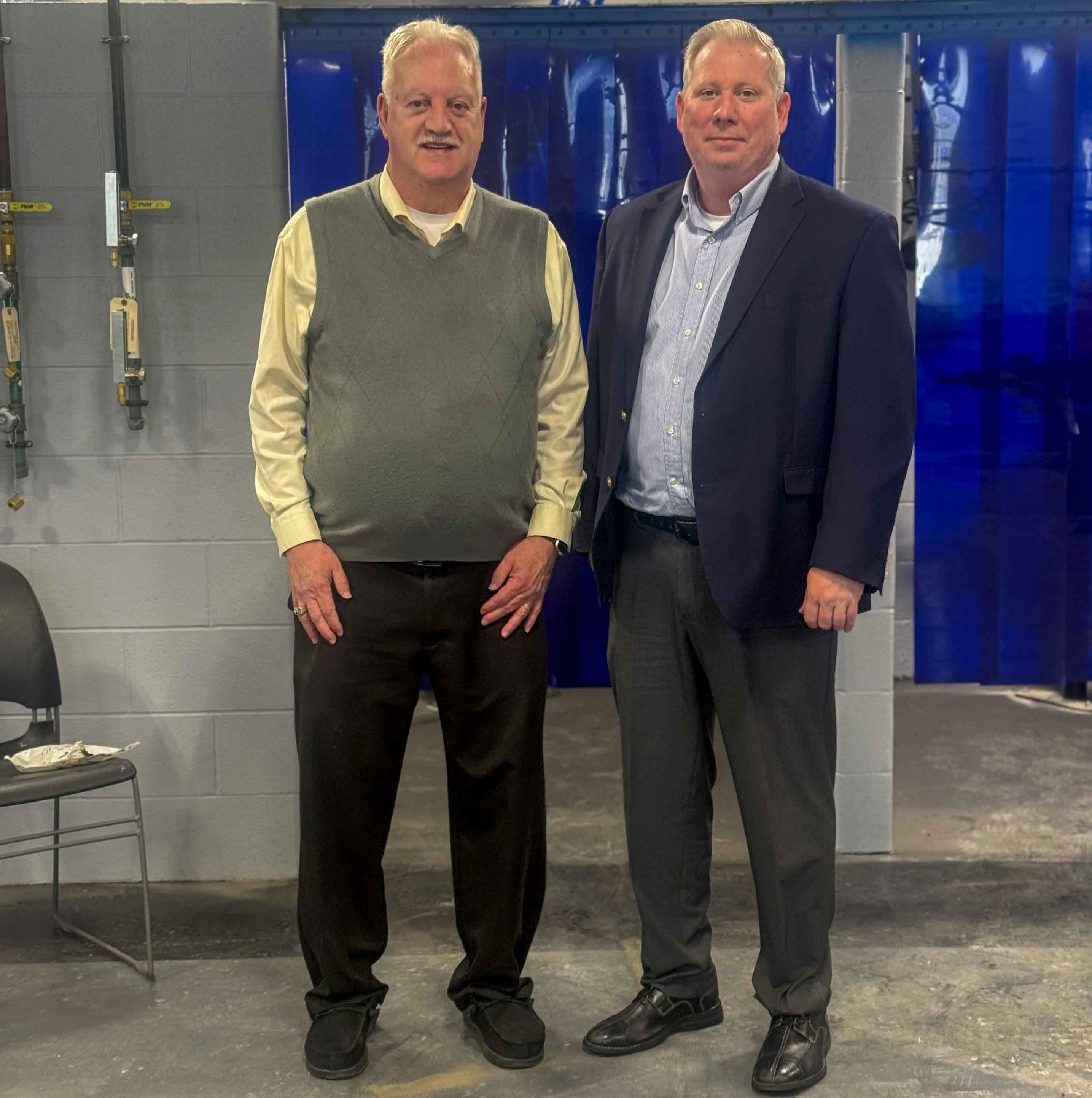 Two men standing side by side in an industrial setting with gray brick walls and blue curtains in the background. The man on the left is wearing a light yellow shirt, gray vest, dark pants, and black shoes. The man on the right is dressed in a blue suit jacket, light blue shirt, dark pants, and black shoes. A black chair with some papers on it is visible to the left