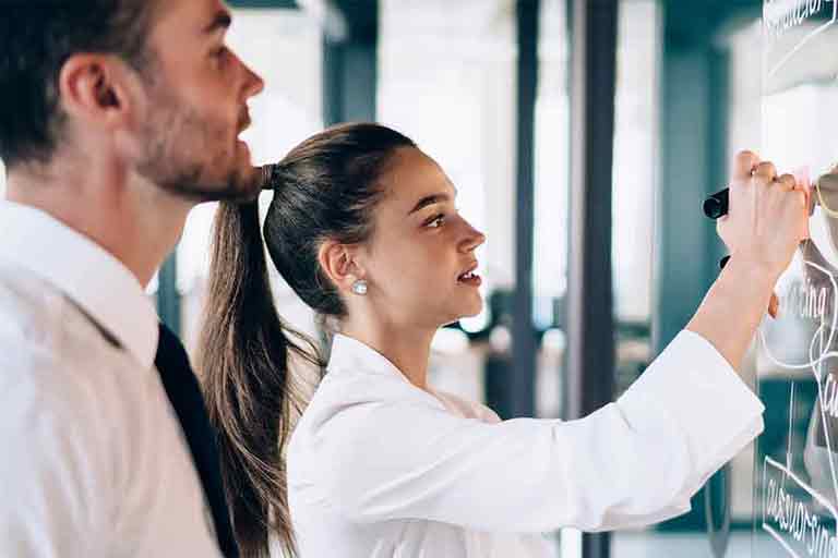 A woman is writing on a glass board with a marker while a man stands beside her observing. Both are dressed in business or professional attire, and they appear to be collaborating in a modern office or meeting space.