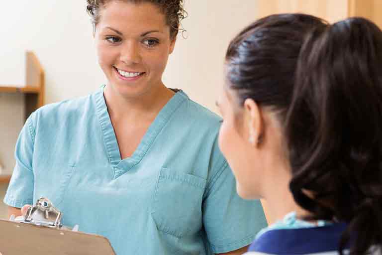 A healthcare professional wearing light blue scrubs is smiling and holding a clipboard while talking to a patient. The patient, with dark hair tied back, is facing the nurse and listening attentively. They appear to be in a medical or clinical setting.