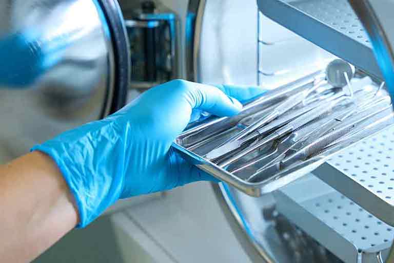 A gloved hand is placing or removing a metal tray of dental or surgical instruments from a sterilization machine, likely an autoclave. The scene suggests a medical or dental setting focused on instrument hygiene and safety.
